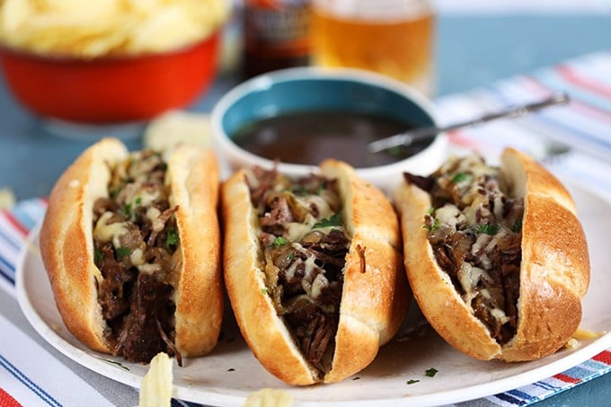 Three Slow Cooker French Dip Sandwiches arranged in a row on a platter with a bowl of au jus in the back
