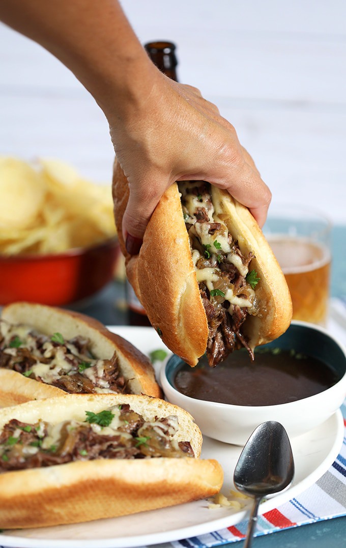 Slow Cooker French Dip Sandwich being dipped into a bowl of au jus on a blue background from TheSuburbansoapbox.com