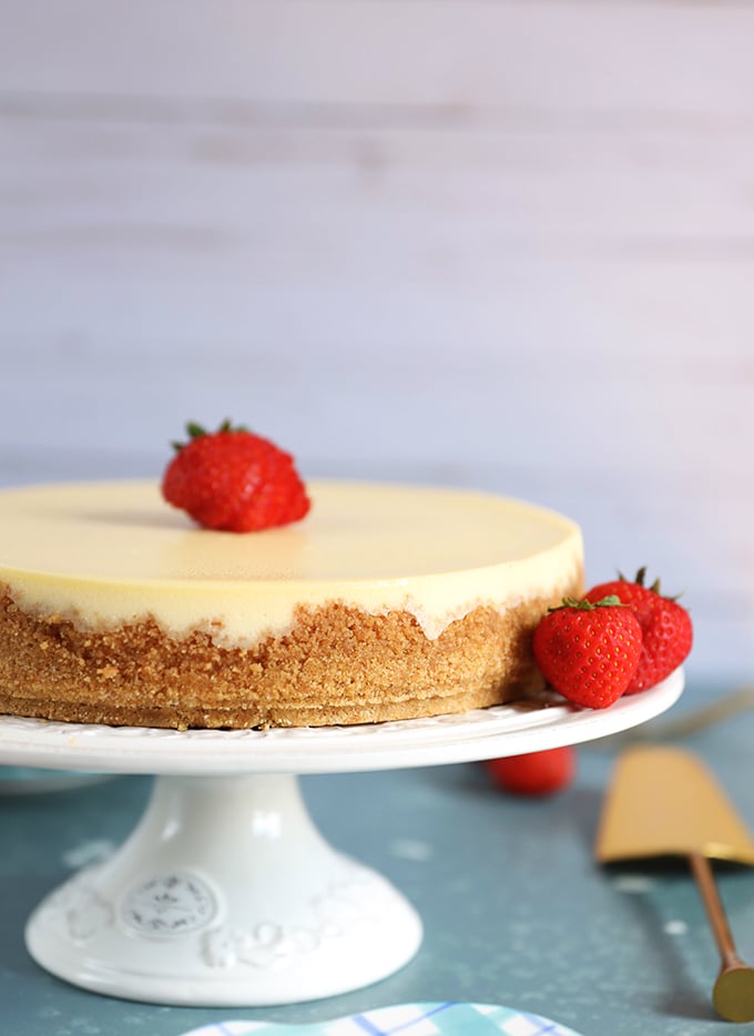 Whole New York Cheesecake on a white cake plate on a blue background.