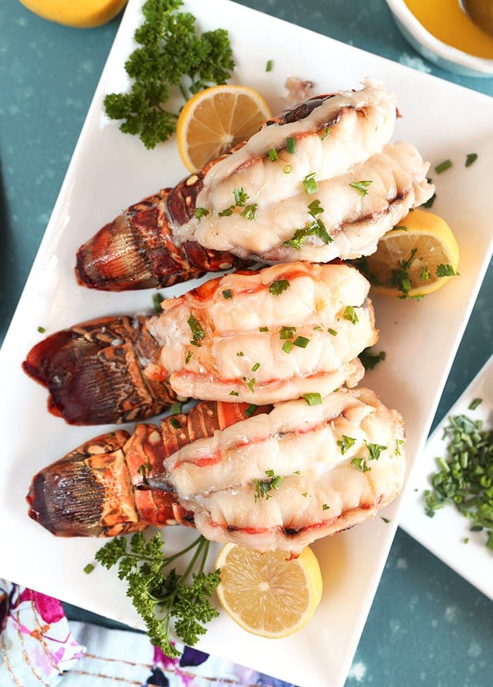 Overhead shot of three baked Lobster Tails with lemons and parsley on a white platter.