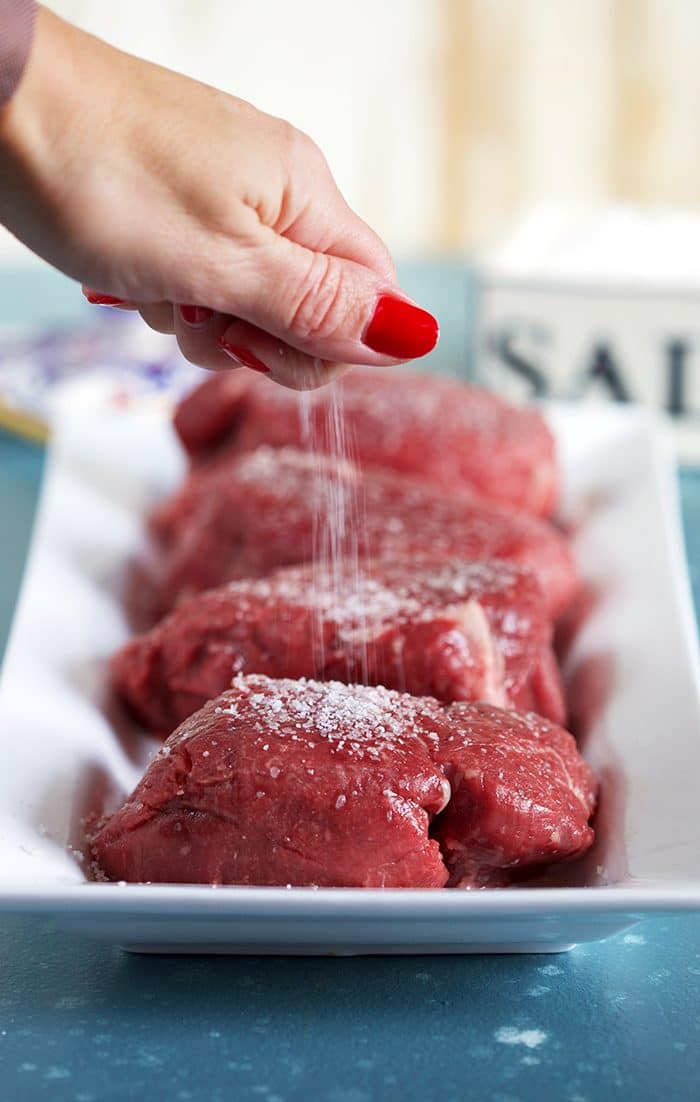 Raw filet mignon on a white plater with kosher salt being sprinkled over them.