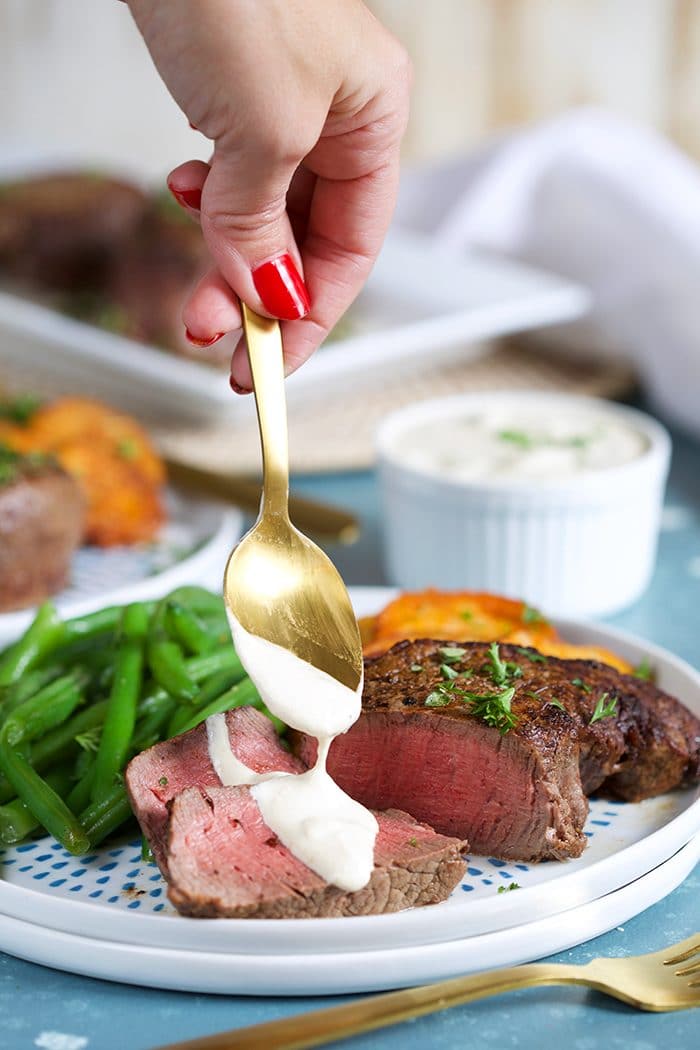 Filet Mignon with horseradish sauce being spooned over the sliced steak on a white plate with green beans.