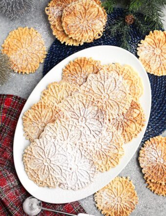 Overhead shot of platter of pizzelles on a gray background with bottle brush trees and a plaid napkin.