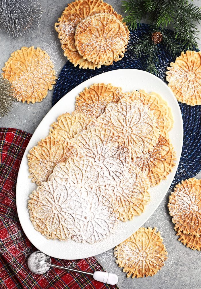 Overhead shot of platter of pizzelles on a gray background with bottle brush trees and a plaid napkin.