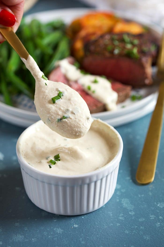Creamy Horseradish Sauce on a gold spoon over a bowl of horseradish sauce on a blue background.