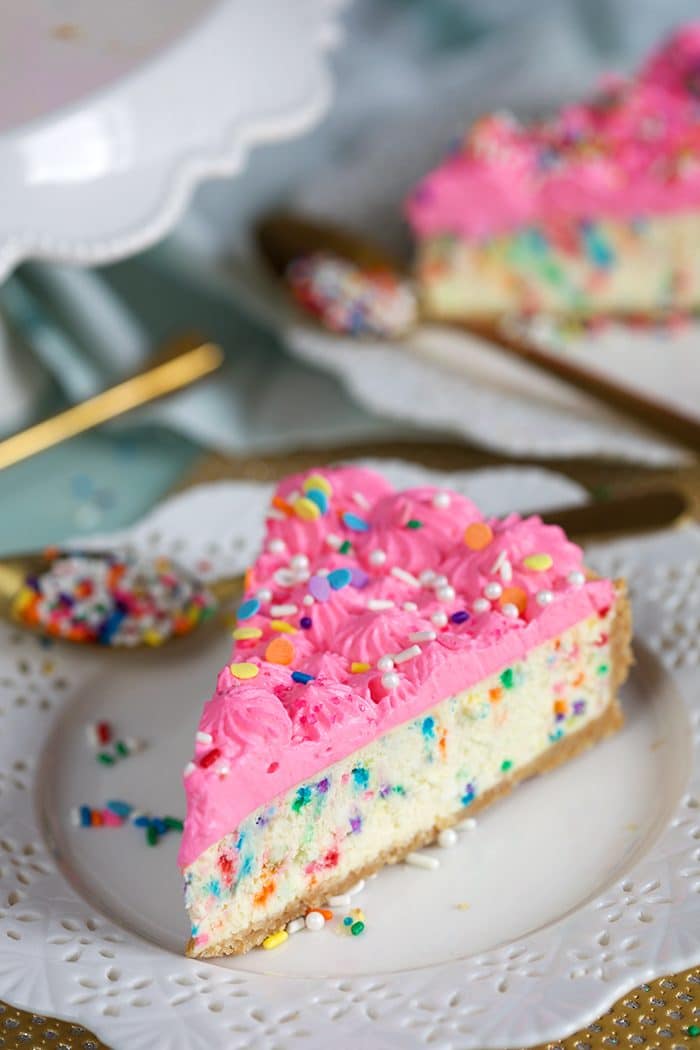 Slice of Funfetti Cheesecake on a white plate with a gold spoon filled with rainbow sprinkles.