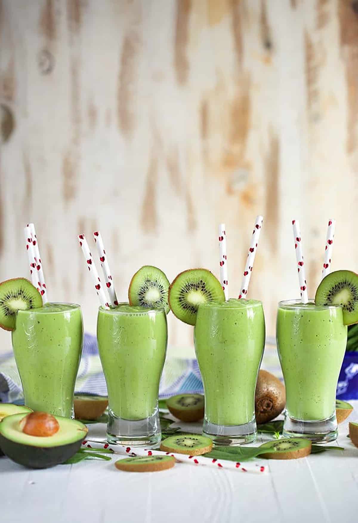 Four kiwi Pineapple Spinach Smoothies in a row with white straws with hearts on them.