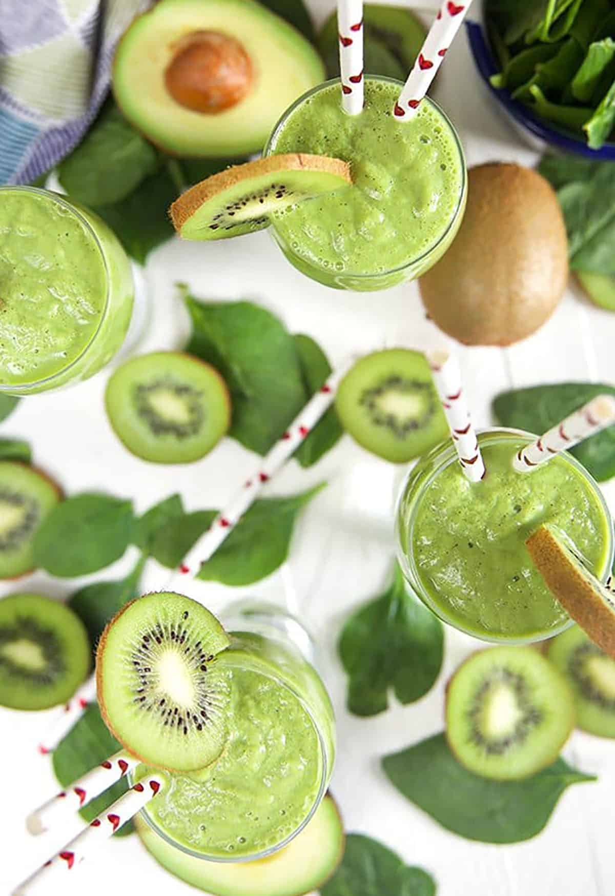 Overhead shot of three Kiwi Pineapple Spinach Smoothies on a white background with kiwi slices and spinach leaves around.