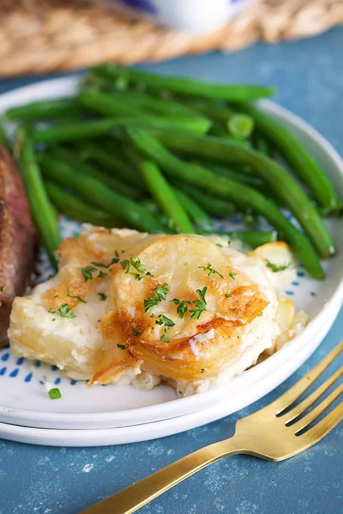 Close up of homemade scalloped potatoes on a white plate with green beans and a gold fork on a blue background.