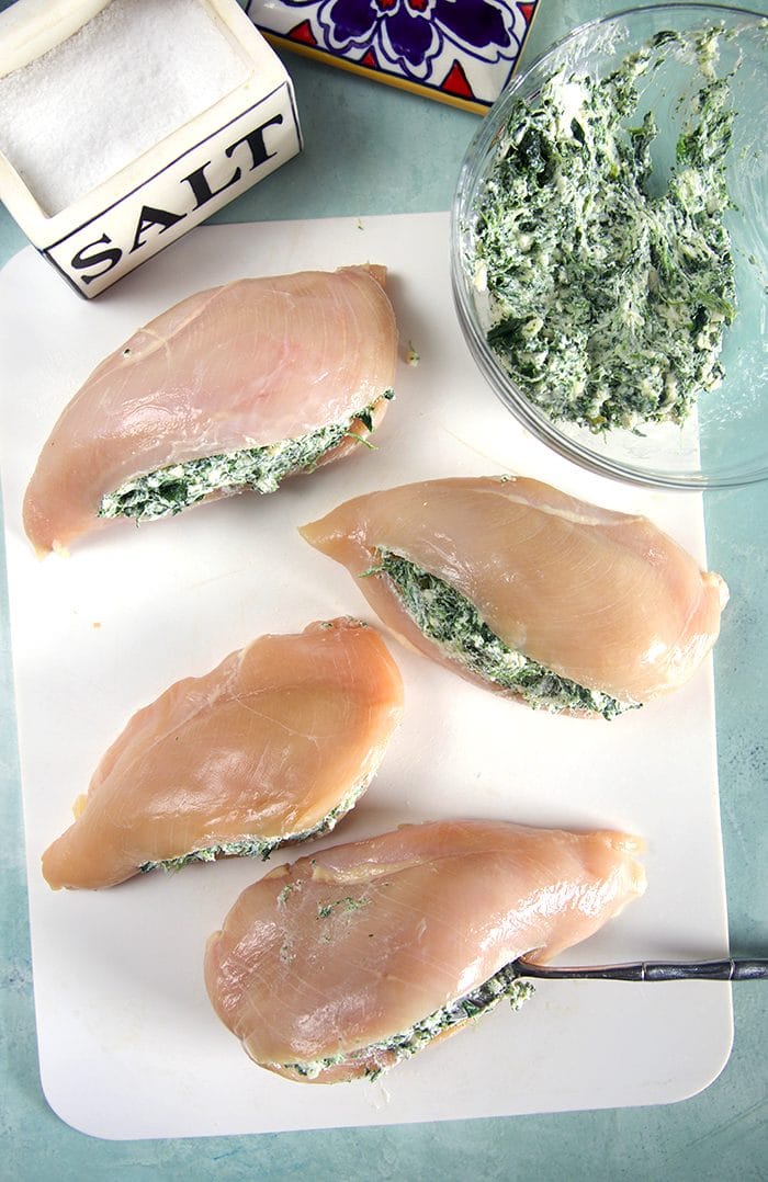 Overhead shot of four raw chicken breasts with spinach dip stuffing and a salt box on a white cutting board.
