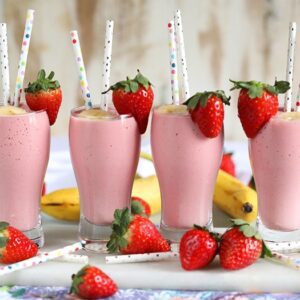Four Strawberry Banana Smoothies in pilsner glasses with paper strawberry and a strawberry on the side.