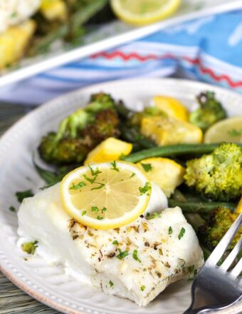 Lemon Baked Cod with a lemon slice on top and a pile of green veggies on a white plate with a fork.