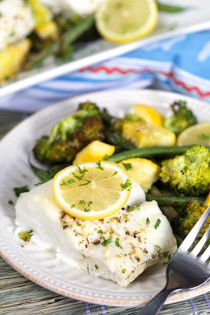 Lemon Baked Cod with a lemon slice on top and a pile of green veggies on a white plate with a fork.