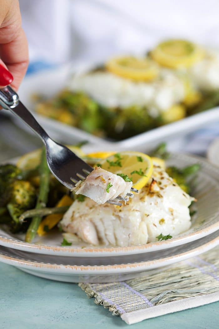A fork with a bite of lemon baked cod on a white plate with green vegetables.