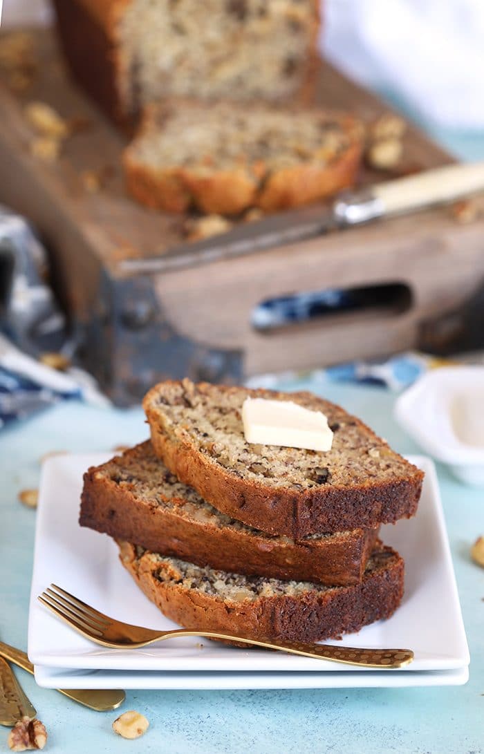 Three sliced of banana nut bread with a pat of butter on top on a white square plate with a gold fork.