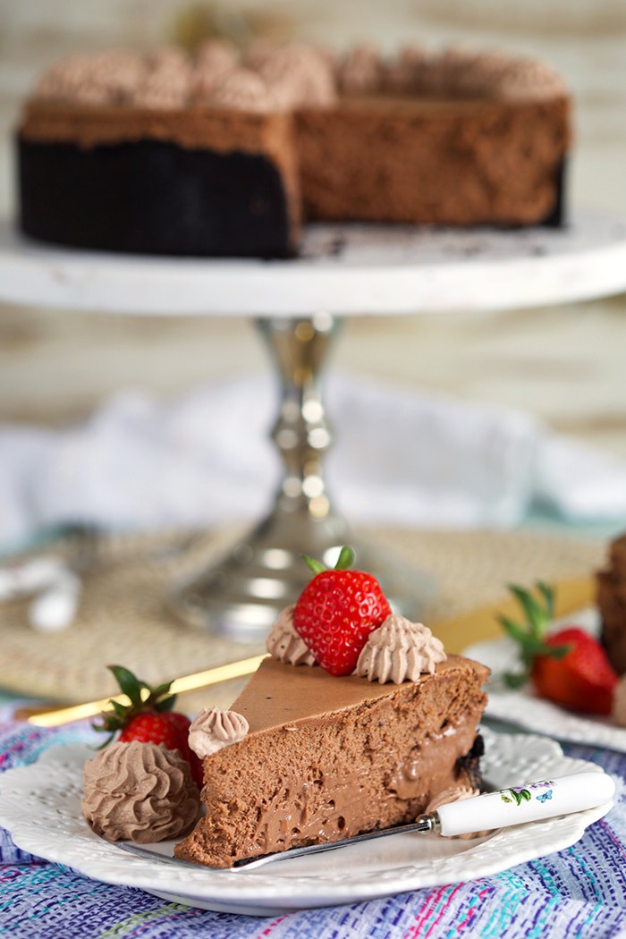 Slice of chocolate cheesecake with a strawberry on top on a white plate with a whole chocolate cheesecake on a marble cake plate in the background.