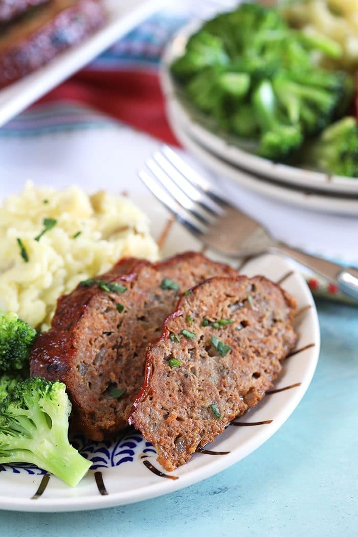 Two slices of crockpot meatloaf on a white plate with mashed potatoes and broccoli.