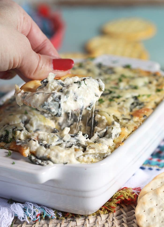Cracker being dipped into hot Spinach Artichoke Dip in a white dish.