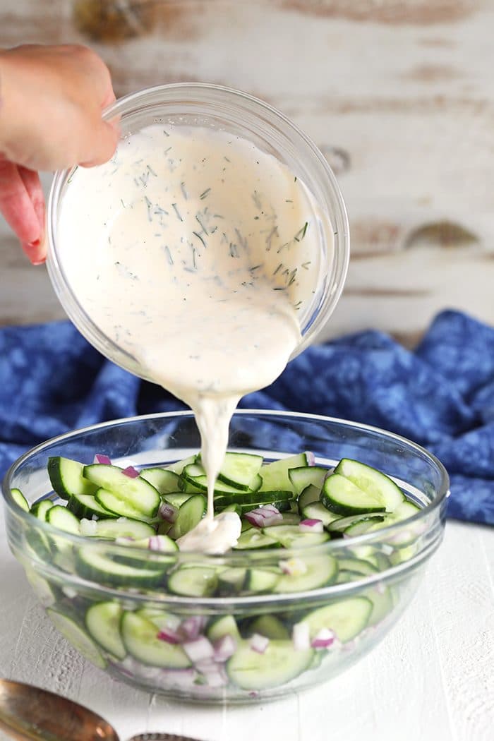 Creamy Cucumber Salad Dressing being poured over cucumbers in a glass bowl.