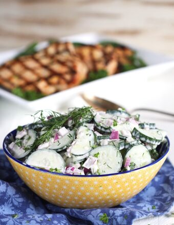 Creamy Cucumber Salad with red onion and dill in a yellow bowl on a blue napkin.