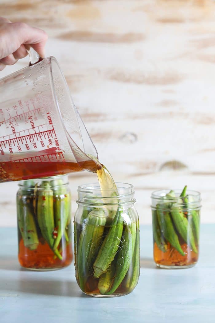 Okra in canning jars with a measuring cup pouring brine into the jars.