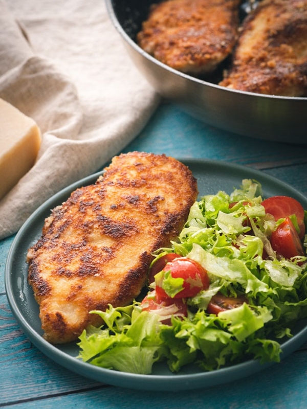 Parmesan CRusted Chicken Cutlet on a plate with a green salad on a blue background.
