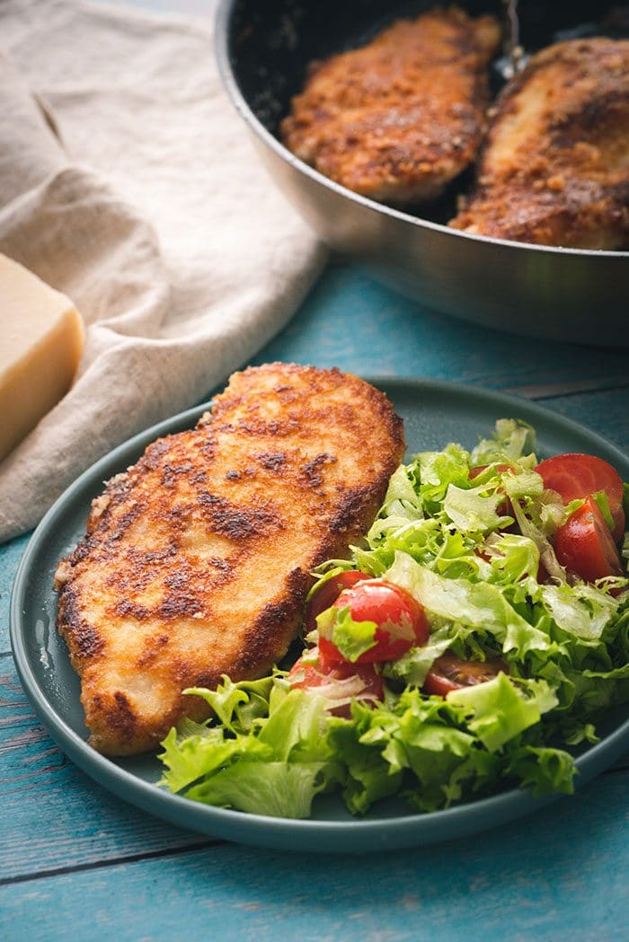 Parmesan CRusted Chicken Cutlet on a plate with a green salad on a blue background.