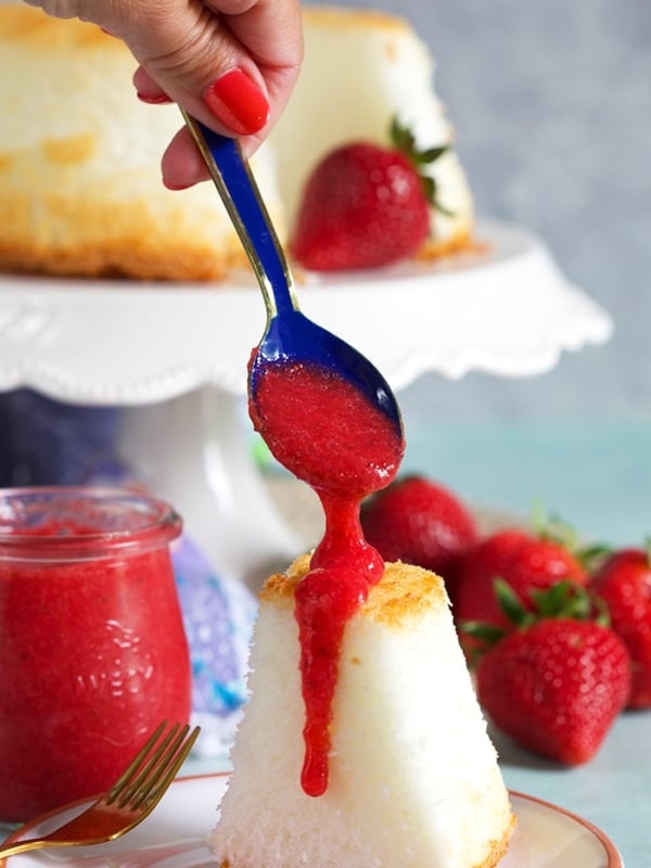 Blue spoon drizzling strawberry puree over an angel food cake on a white plate.