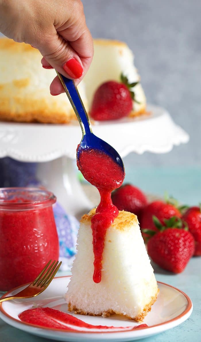 Blue spoon drizzling strawberry puree over an angel food cake on a white plate.