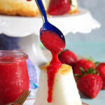 Blue spoon drizzling strawberry puree over an angel food cake on a white plate.