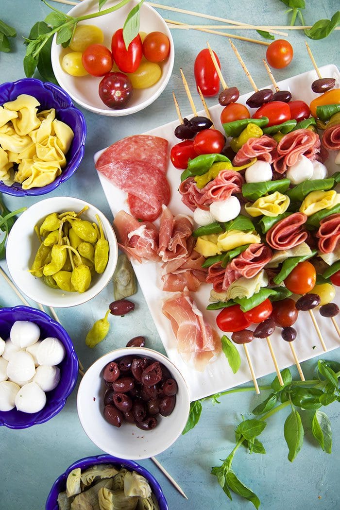 Antipasto skewers on a white platter being assembles with ingredients in the blue bowls.