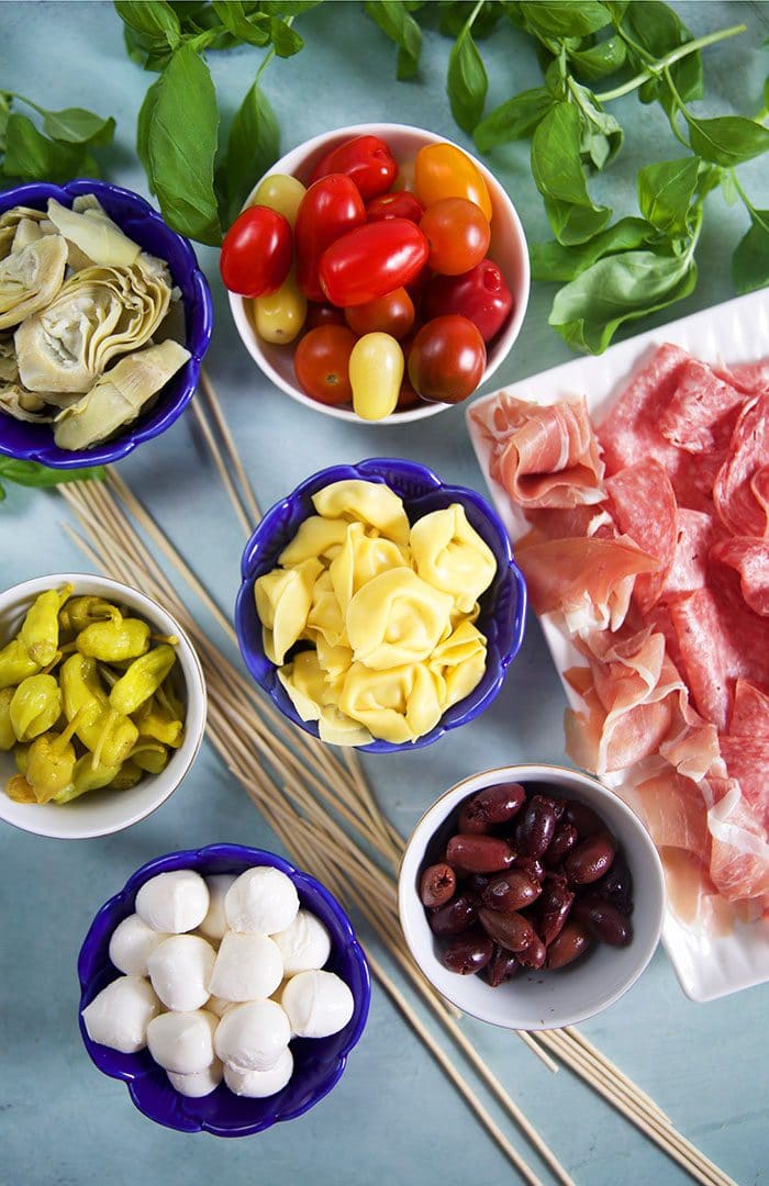 Ingredients for antipasto skewers in blue bowls and charcuterie on a platter.