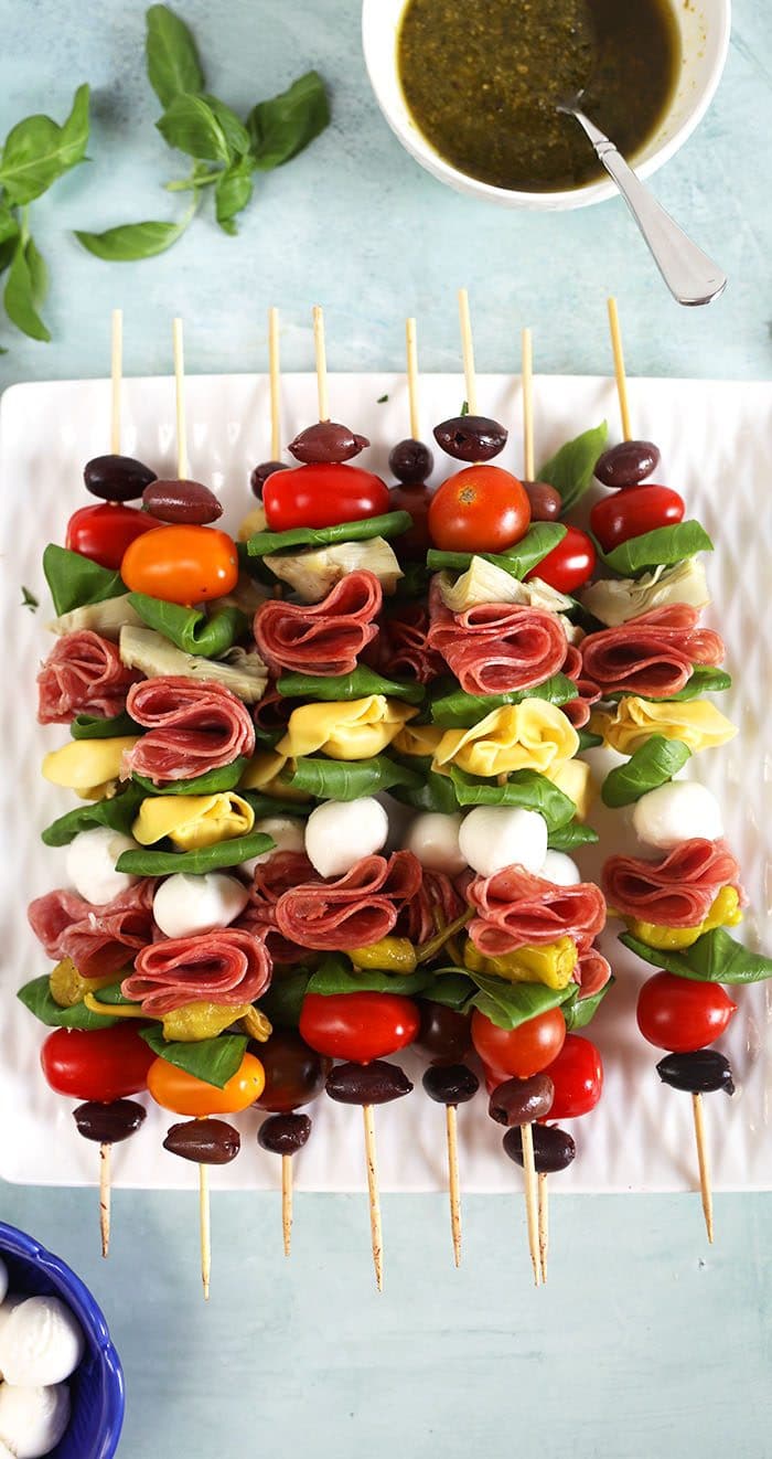 Overhead shot of Antipasto skewers on a white square plate with a bowl of pesto in the upper right corner.