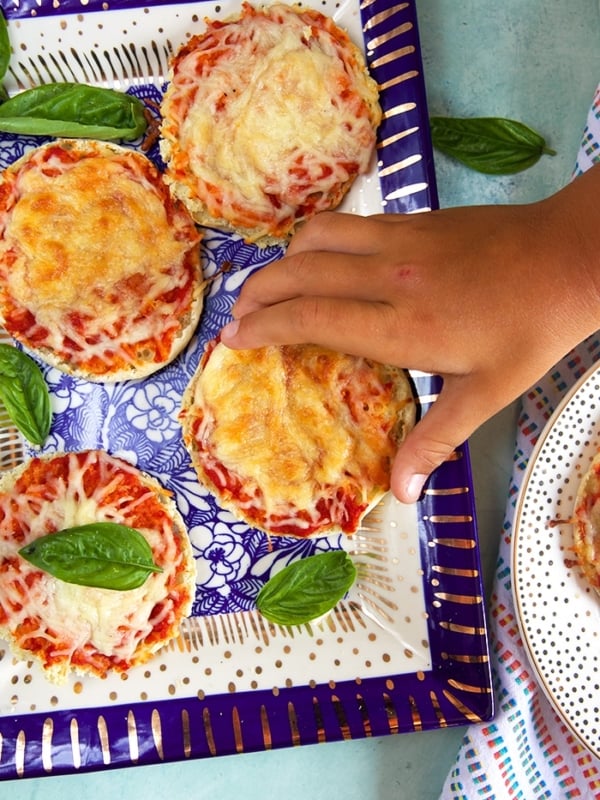 Child's hand taking an english muffin pizza off a blue and white platter.