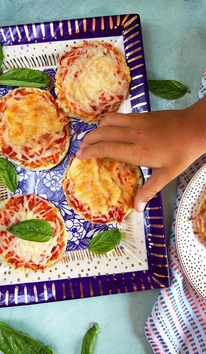 Child's hand taking an english muffin pizza off a blue and white platter.