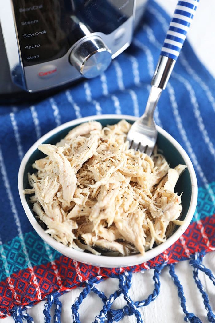 Shredded chicken breast in a white bowl with a fork on a blue and white striped napkin.