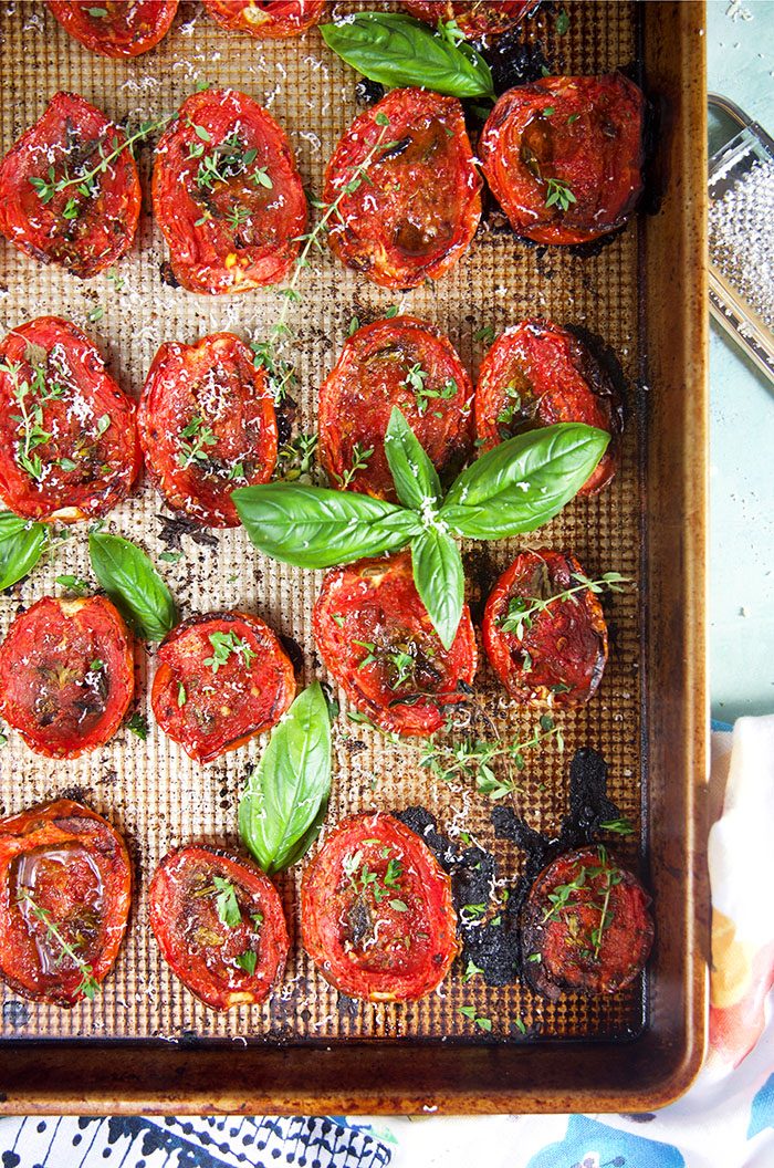 Overhead shot of roasted tomatoes on a baking sheet with basil and herbs.
