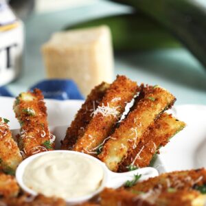 Zucchini Fries on a white platter with a wedge of parmesan and zucchini in the background.
