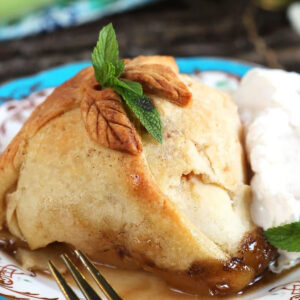 Apple dumpling topped with a sprig of mint on a blue and white plate with whipped cream and a gold fork with a white handle.