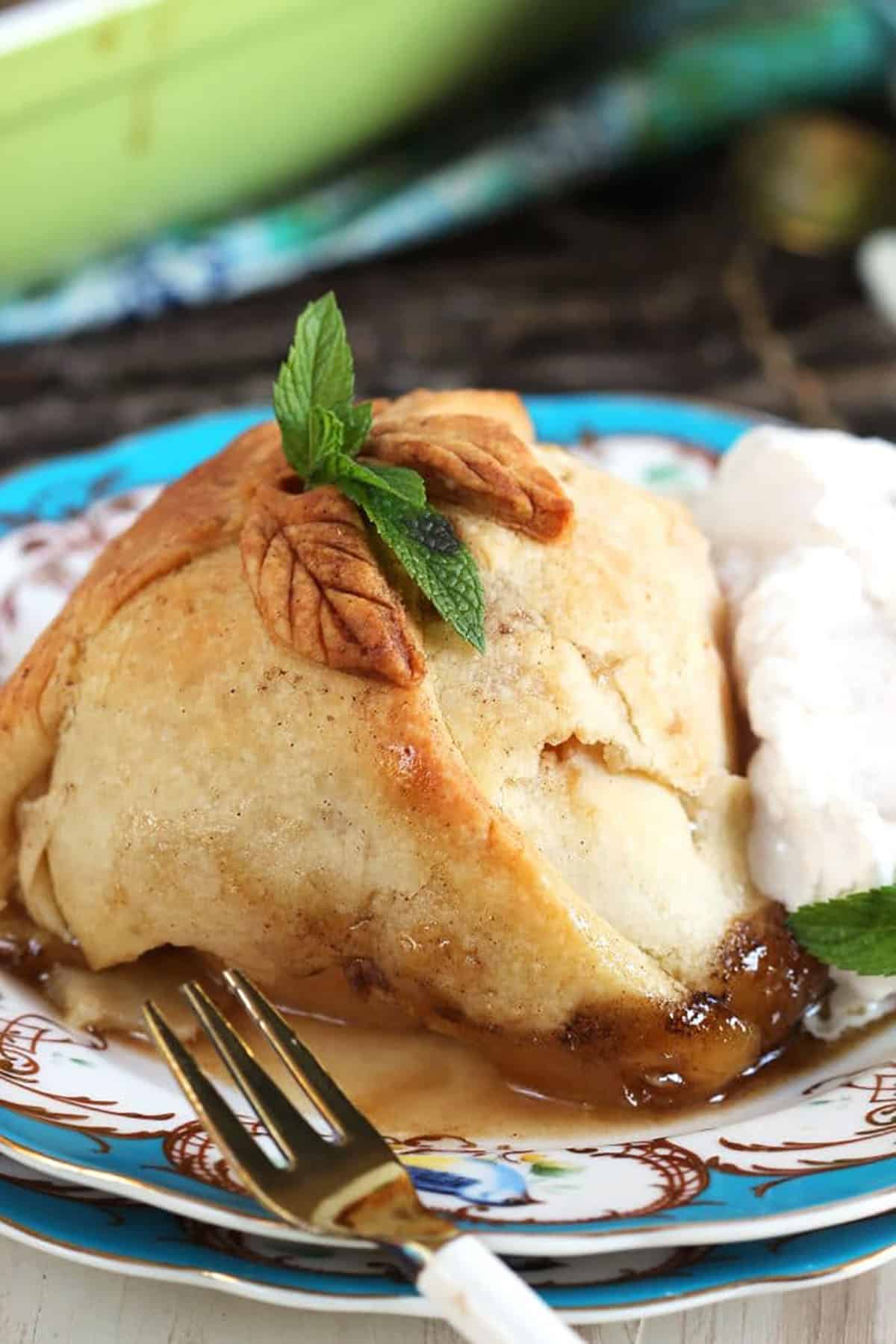 Apple dumpling topped with a sprig of mint on a blue and white plate with whipped cream and a gold fork with a white handle.