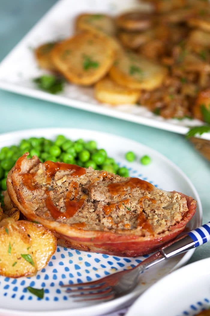 Slice of bacon wrapped meatloaf on a white plate with peas.