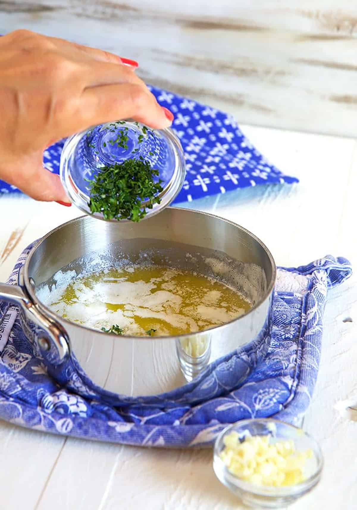 Melted butter in a skillet on a blue pot holder with herbs being poured into the butter.