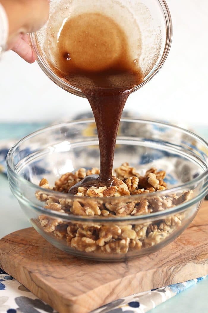 Brown sugar syrup being poured over walnuts in a glass bowl.