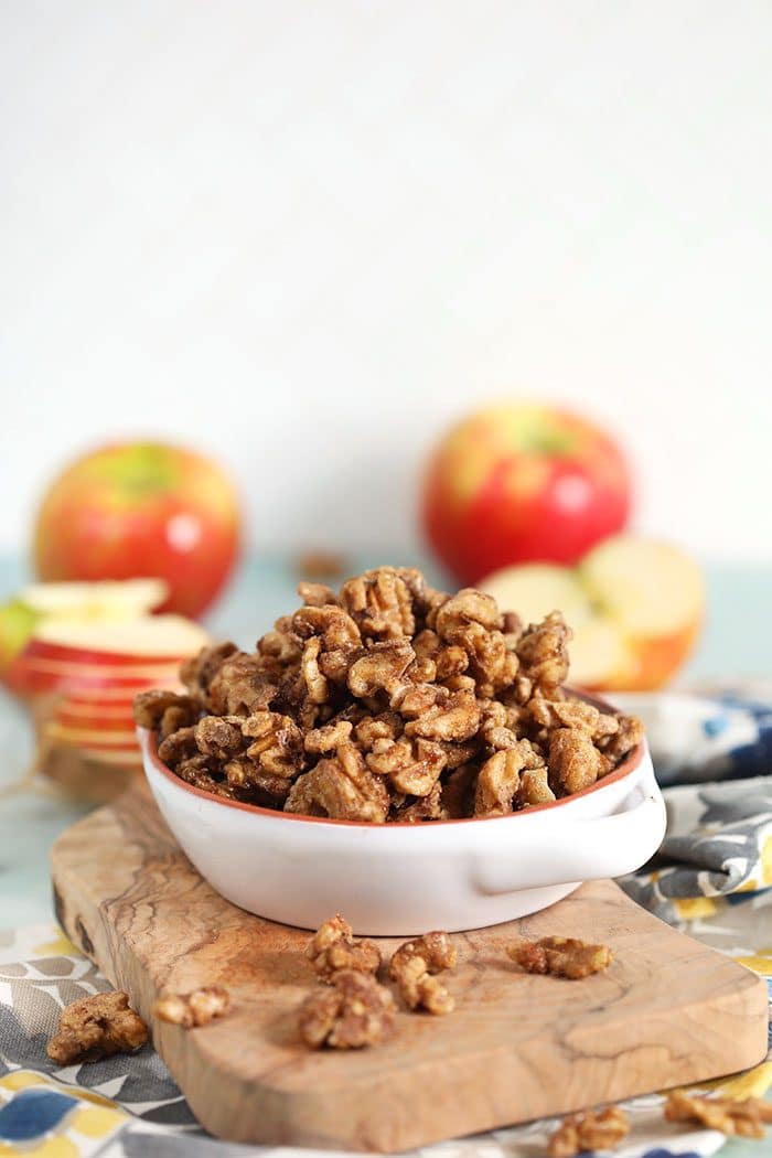 Candied Walnuts in a white bowl on a wood board with apples in the background.