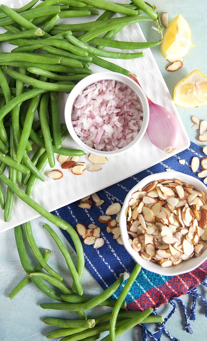 Overhead shot of green beans, chopped shallots, sliced almonds and lemon on a blue background.