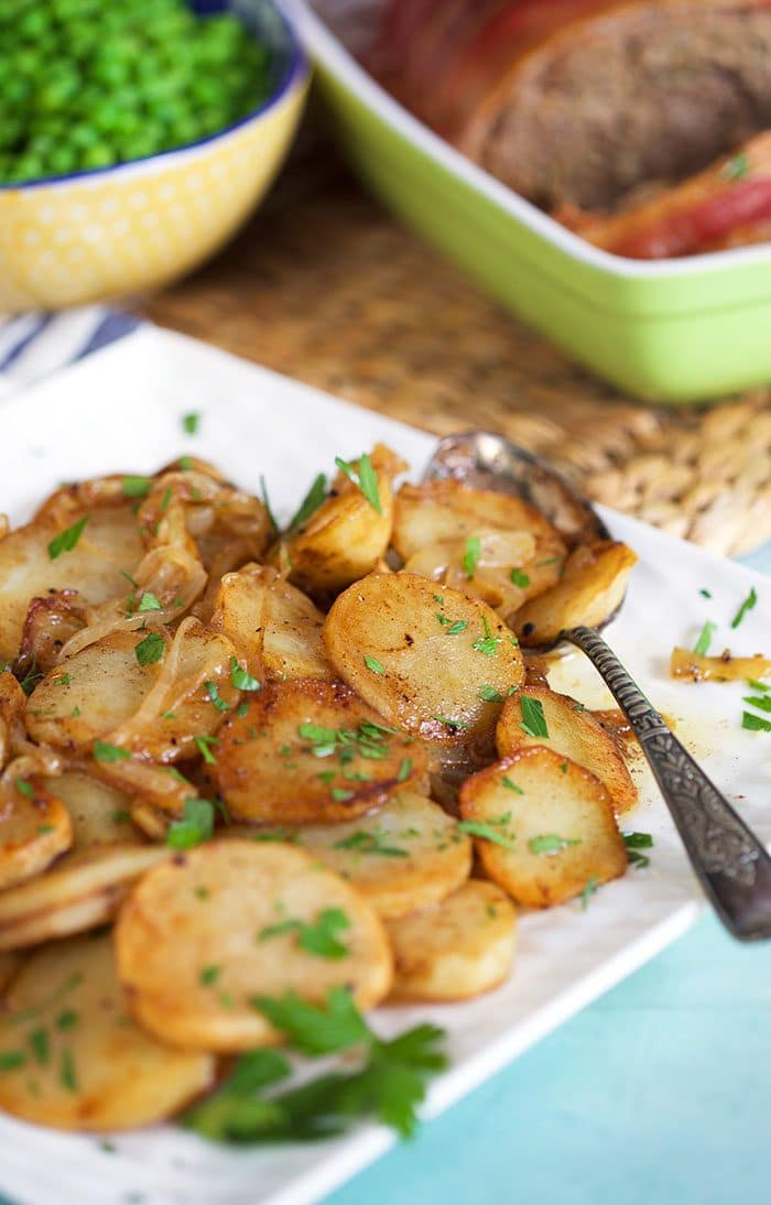 Lyonnaise potatoes on a white platter with a spoon and peas in the background.