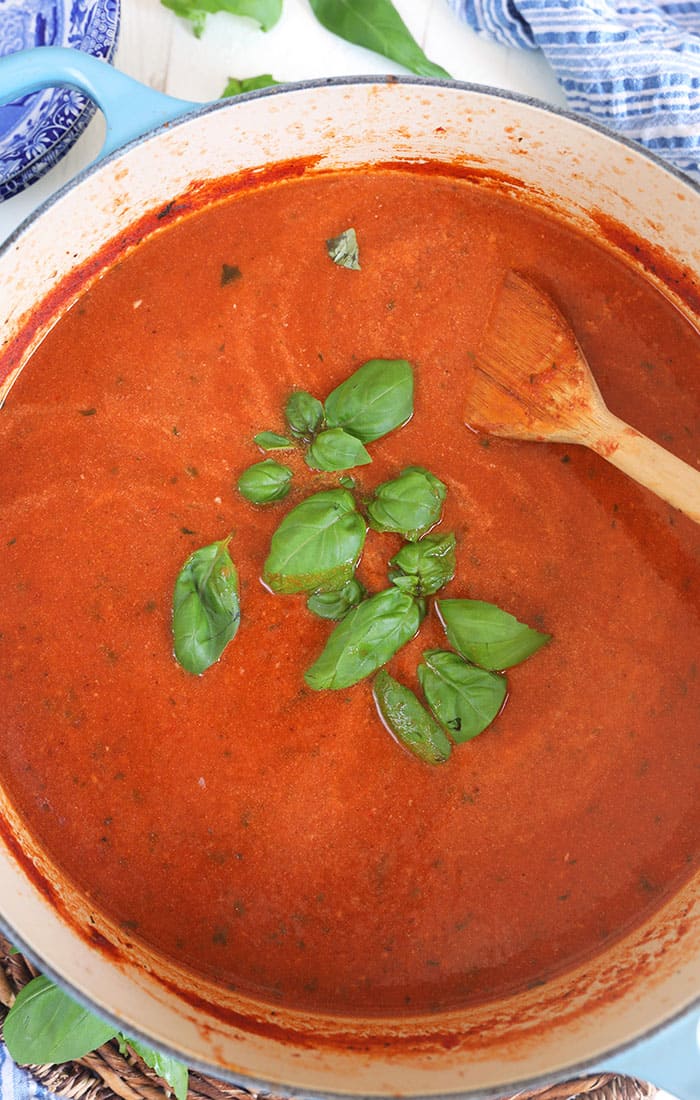 Close up of tomato basil bisque in a pot with a wooden spoon and basil leaves in the middle of the soup.