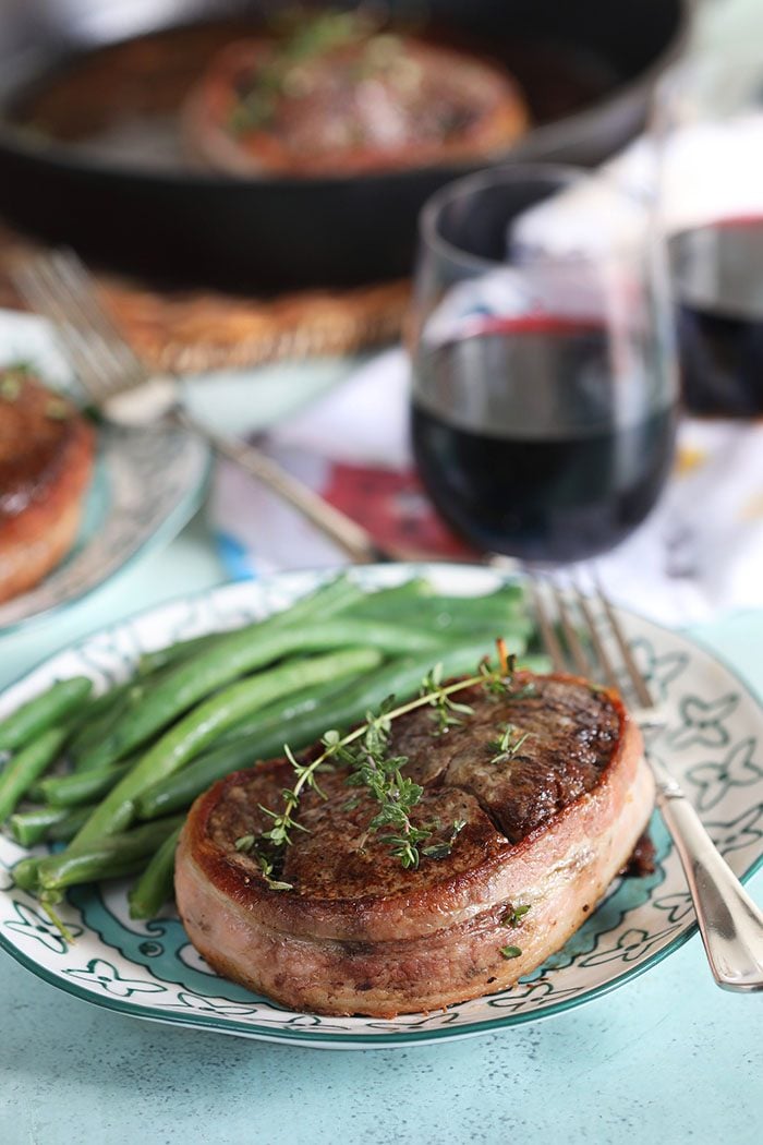 Bacon wrapped filet mignon on a plate with green beans and a glass of wine in the background.