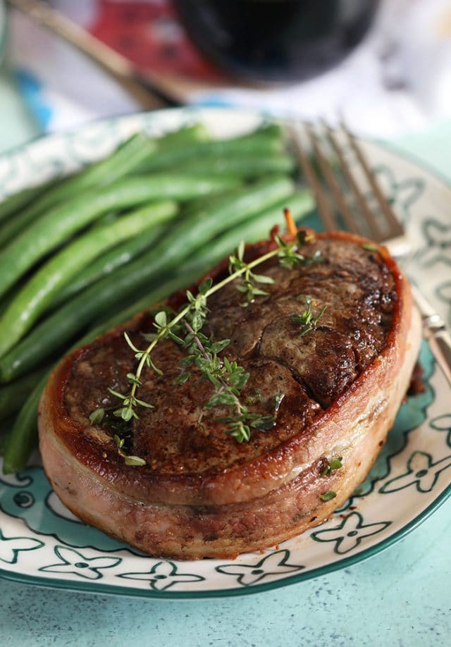 Bacon wrapped filet mignon with thyme sprigs on top and green beans on the plate.