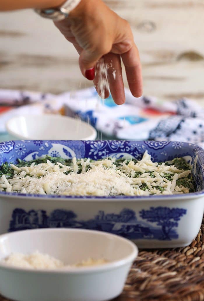 Cheese being sprinkled over creamed spinach in a baking dish.
